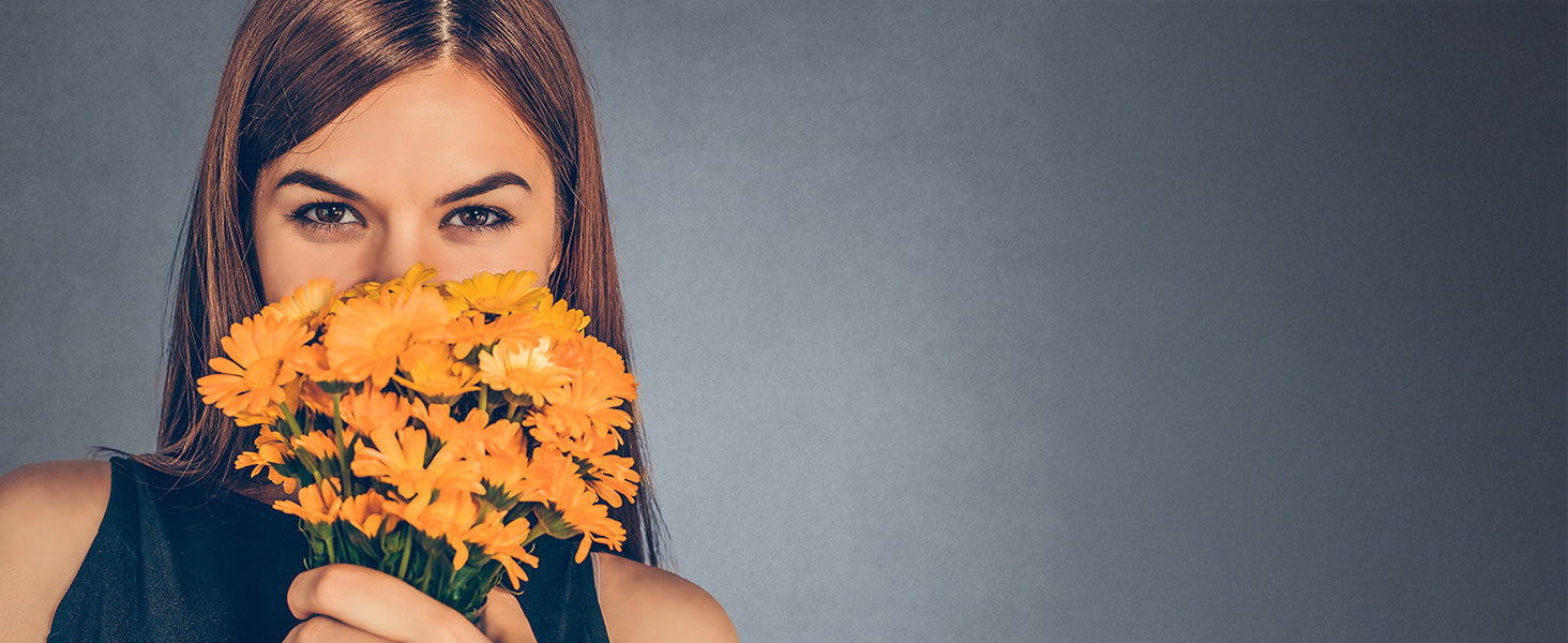 Eine Frau mit glattem braunem Haar hält einen Strauß leuchtend orangefarbener Blumen dicht vor ihr Gesicht, wobei nur ihre Augen zu sehen sind, vor einem schlichten grauen Hintergrund.