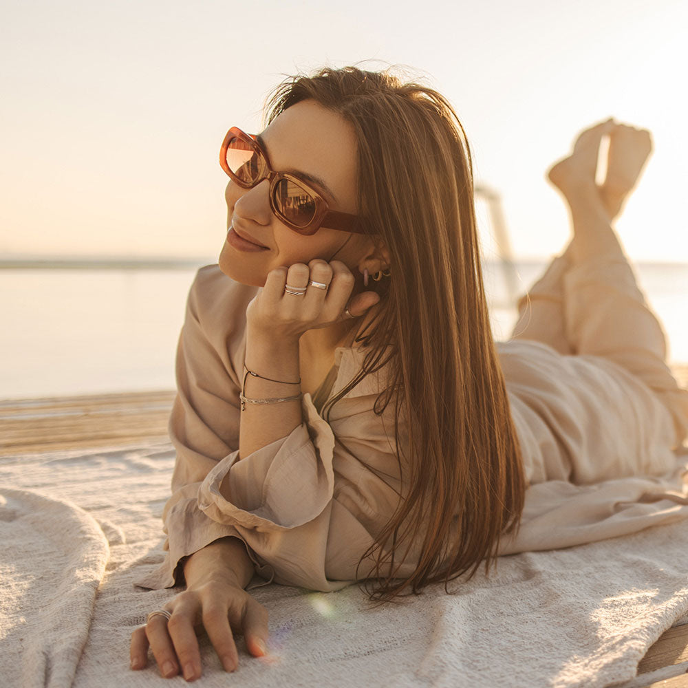 Eine Frau mit Sonnenbrille und heller Kleidung liegt lächelnd im Freien auf einer Decke und stützt ihren Kopf auf ihre Hand. Im Hintergrund sind ein ruhiges Gewässer und sanftes Sonnenlicht zu sehen.