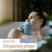 Eine Frau entspannt sich in einem Schaumbad mit einem blauen Becher, umgeben von grünen Pflanzen. Der deutsche Text lautet: Basen-Bad Entspannung pflegen – mit basisch-mineralischem Badesalz von fitne für die Säure-Basen-Balance.
