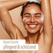 Eine lächelnde Frau mit nassen, seifigen Haaren duscht und hebt die Arme. Auf beigem Hintergrund hebt der Text hervor: fitne Basischer Duschbalsam – pflegend & schützend mit Bio-Aloe Vera und Bio-Mandelöl.