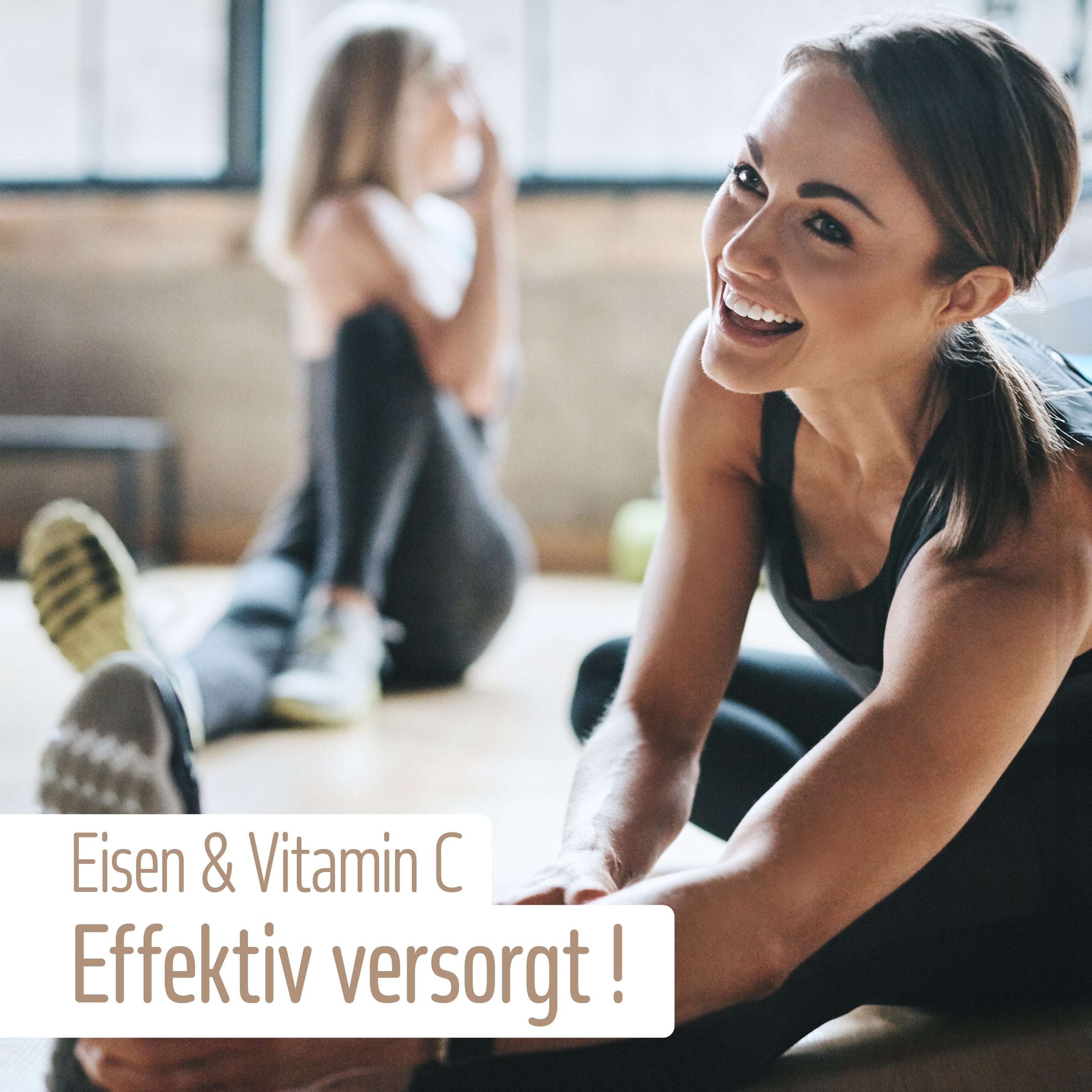 Zwei Frauen in Sportbekleidung dehnen sich in einem hellen Raum auf Yogamatten. Eine lächelt, während sie nach ihrem Fuß greift. Der Text auf Deutsch lautet: Eisen Komplex Kapseln von fitne – Organisch gebundenes Eisen & Vitamin C für effektive Versorgung!.