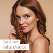 Eine Frau mit langen, welligen roten Haaren und klarer Haut lächelt vor beigem Hintergrund. Der Overlay-Text auf Deutsch lautet: Natürlich schön mit fitne Nährstoffkomplex Haut & Haare – Mikronährstoffe für Ihre Schönheit.