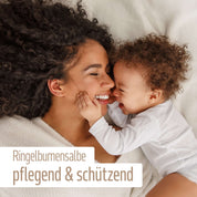 Eine lächelnde Frau und ein glückliches Baby kuscheln auf einem Bett. Text in Deutsch: Mit Bio Olivenöl und Calendula - Bio Ringelblumen Salbe von FITNE Health Care, pflegend & schützend.