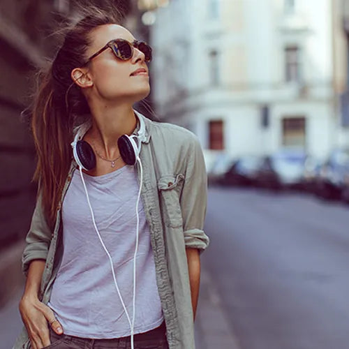 Eine junge Frau mit Sonnenbrille und Kopfhörern geht lächelnd eine Straße entlang. Sie trägt lange, zurückgebundene Haare und ist leger gekleidet, trägt ein Hemd über einem T-Shirt.