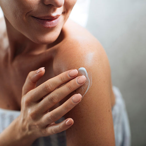 Eine Frau lächelt sanft, während sie mit der Hand eine kleine Menge Lotion auf ihre nackte Schulter aufträgt.