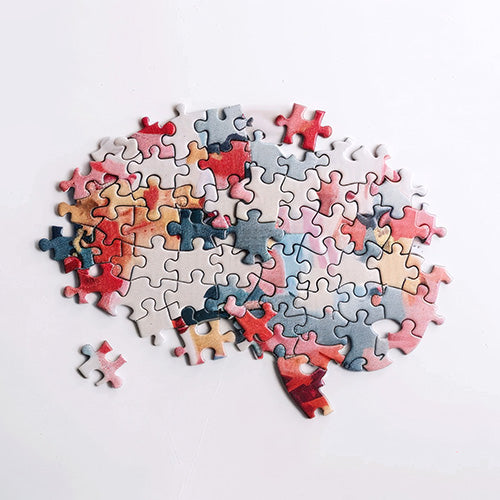 Ein Puzzle in Gehirnform mit mehreren fehlenden Teilen liegt auf einer weißen Fläche. Das Puzzle ist in verschiedenen gedeckten Farben gehalten, darunter Rot-, Blau- und Gelbtöne.