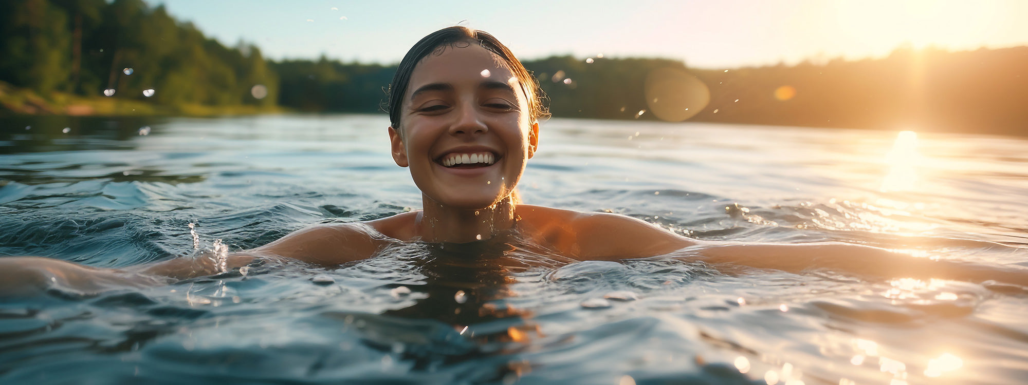Eine junge Frau lächelt fröhlich, während sie bei Sonnenuntergang in einem See schwimmt, um sie herum plätschern Wassertropfen, im Hintergrund sind Bäume zu sehen.