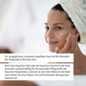 Eine Frau mit einem Handtuch um den Kopf lächelt, während sie fitne Basische Körperlotion aufträgt. Der deutsche Text hebt die Vorteile von Bio-Mandelöl und Bio-Sheabutter für weiche, gepflegte Haut hervor.